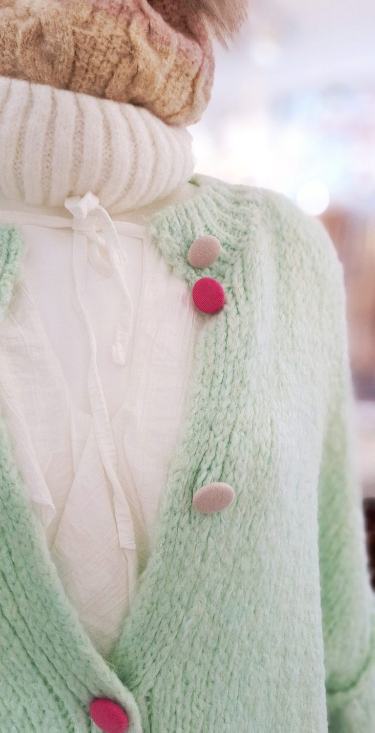 Grüne Strickjacke mit pinken und Knöpfen über weißem Oberteil.